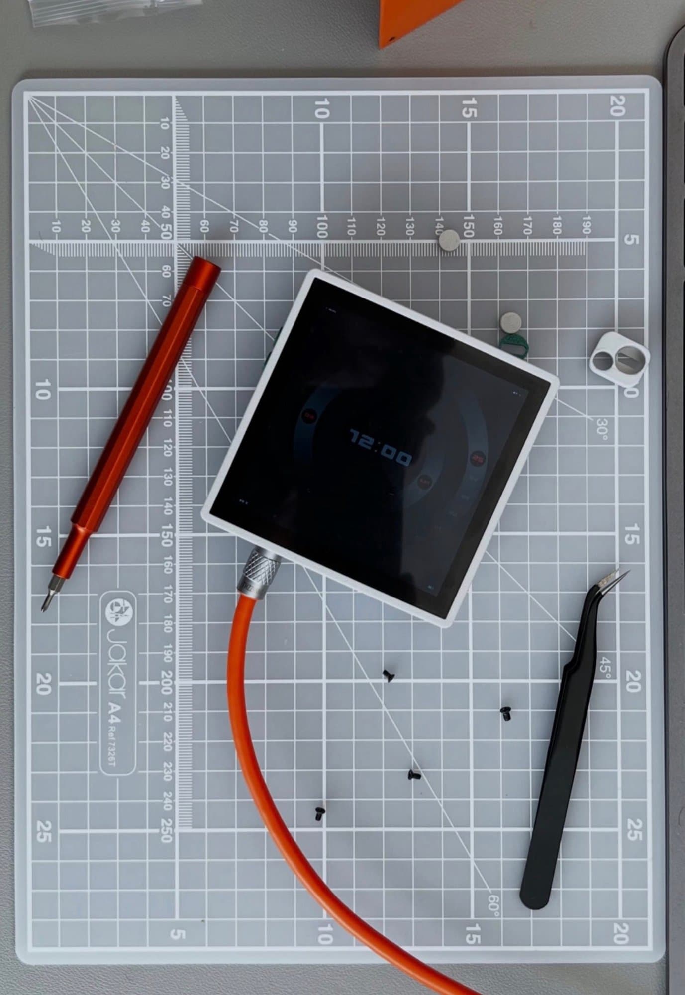 A flat lay photograph of a square digital display module being disassembled or repaired on a grid cutting mat alongside precision tools.