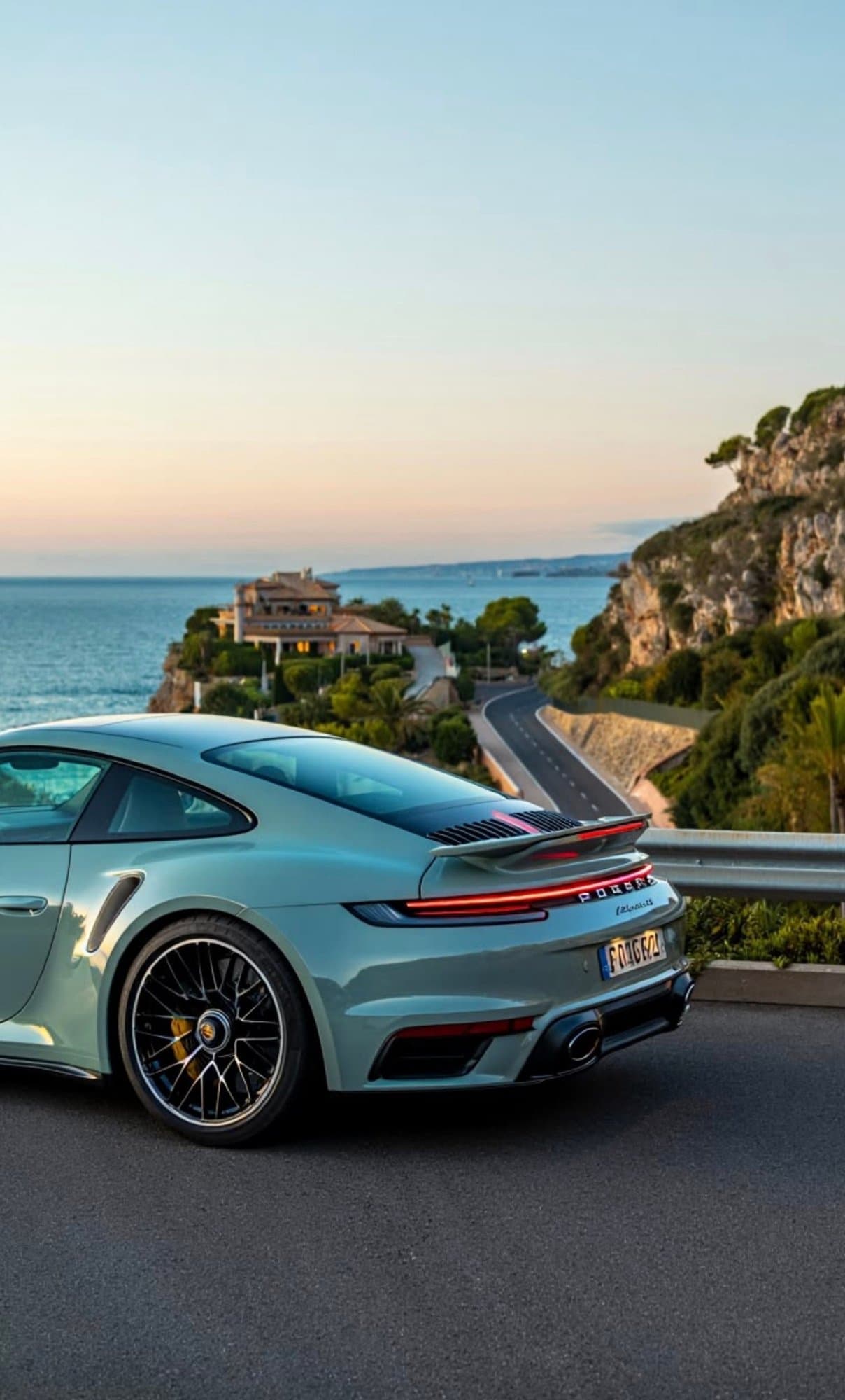 A pale sage green Porsche 911 Turbo S parked along a winding coastal road overlooking the sea at dusk.