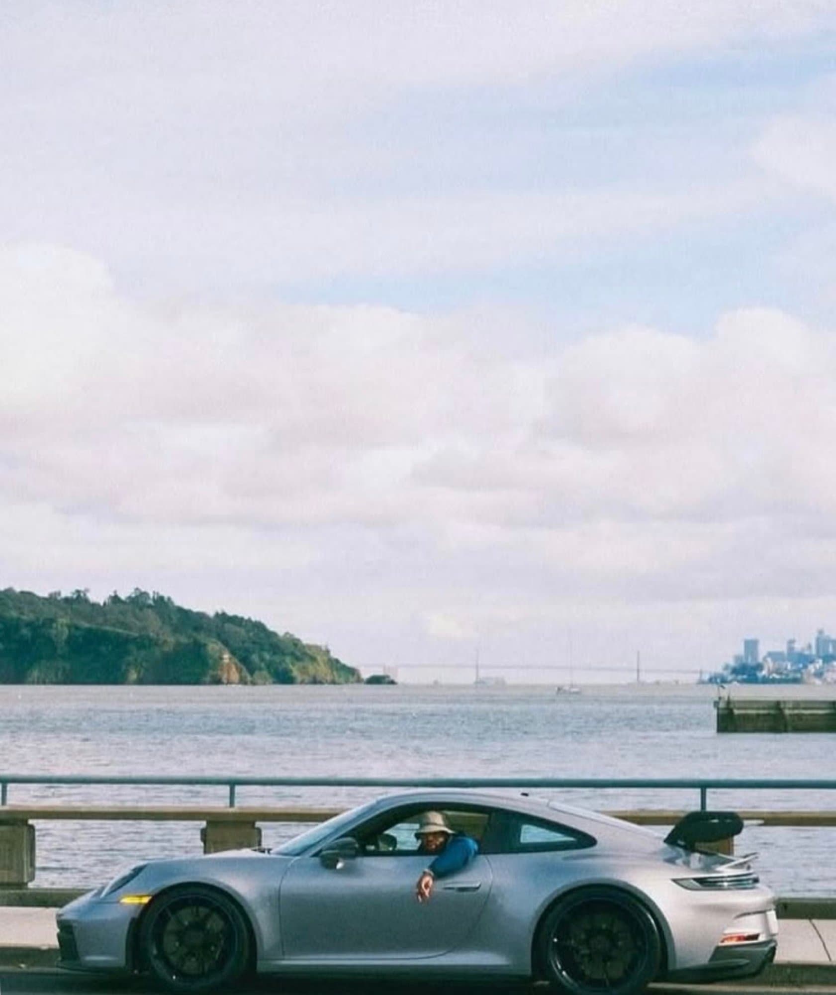 A silver Porsche 911 GT3 (992) parked along a coastal waterfront with the driver resting his arm out the window.