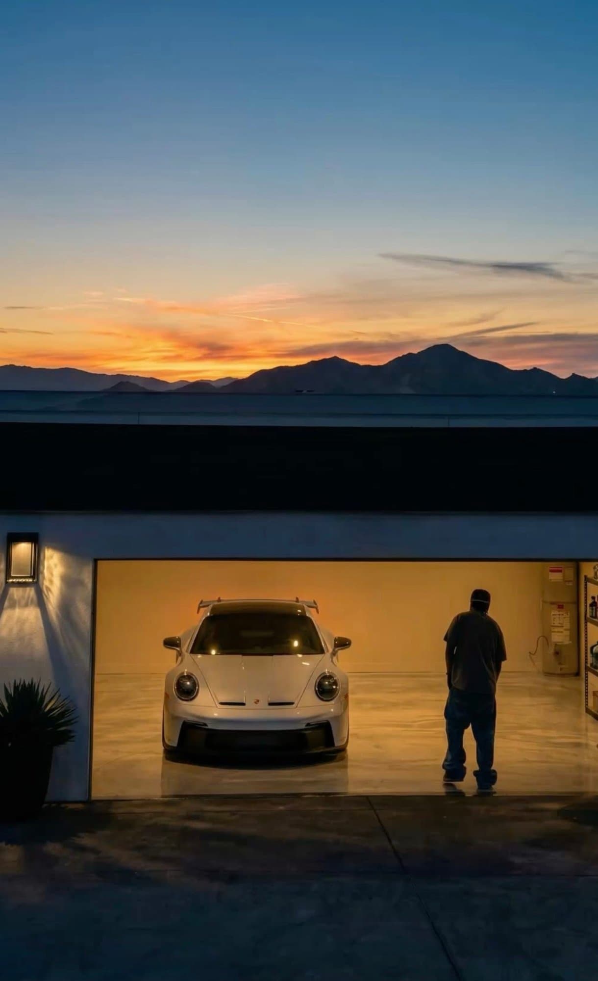 A white Porsche 911 GT3 sits inside a warmly lit garage against a desert sunset framed by mountain silhouettes.