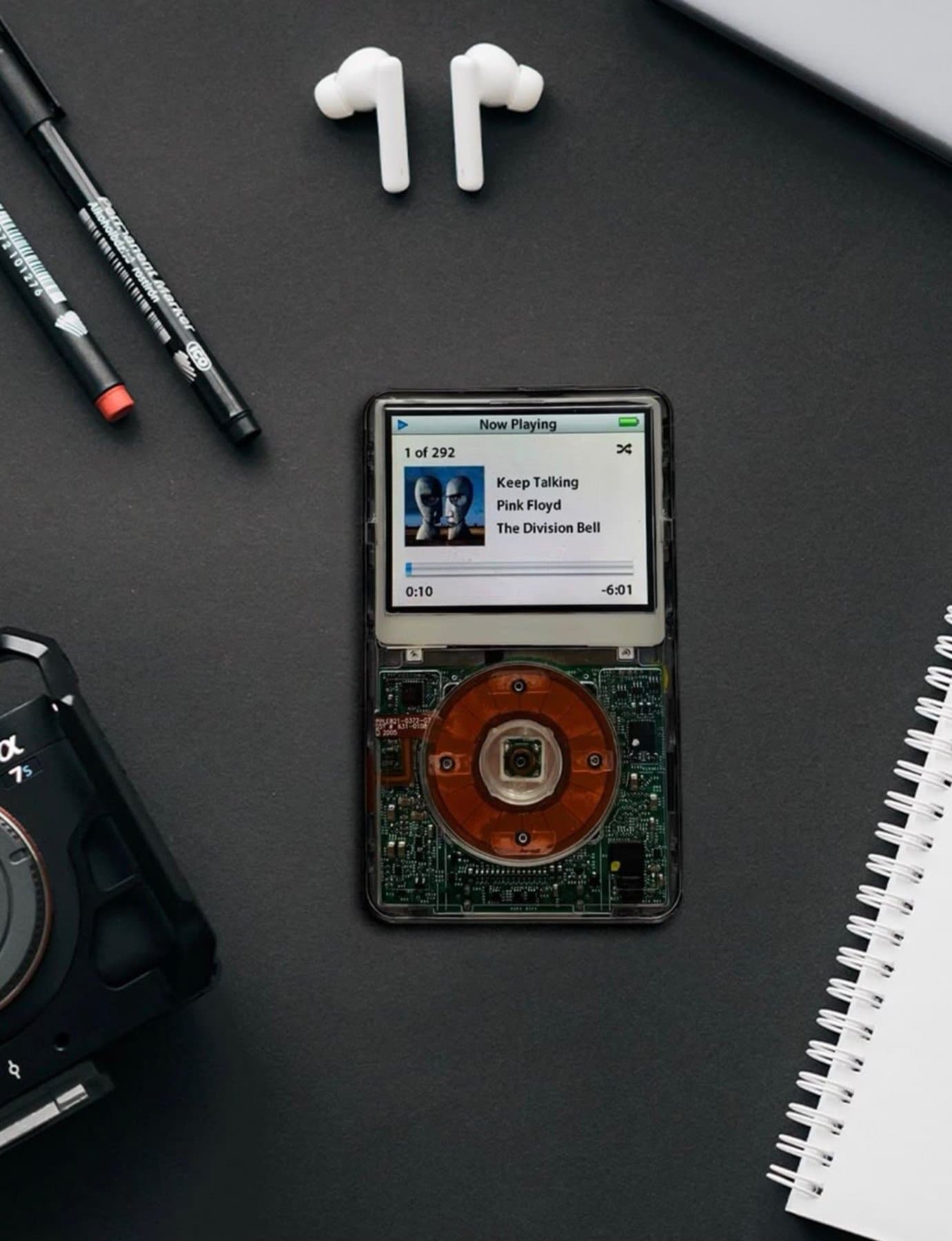 A flat lay featuring a custom transparent-shelled Apple iPod Classic showing internal circuitry, paired with wireless earbuds, pens, and a camera on a black surface.
