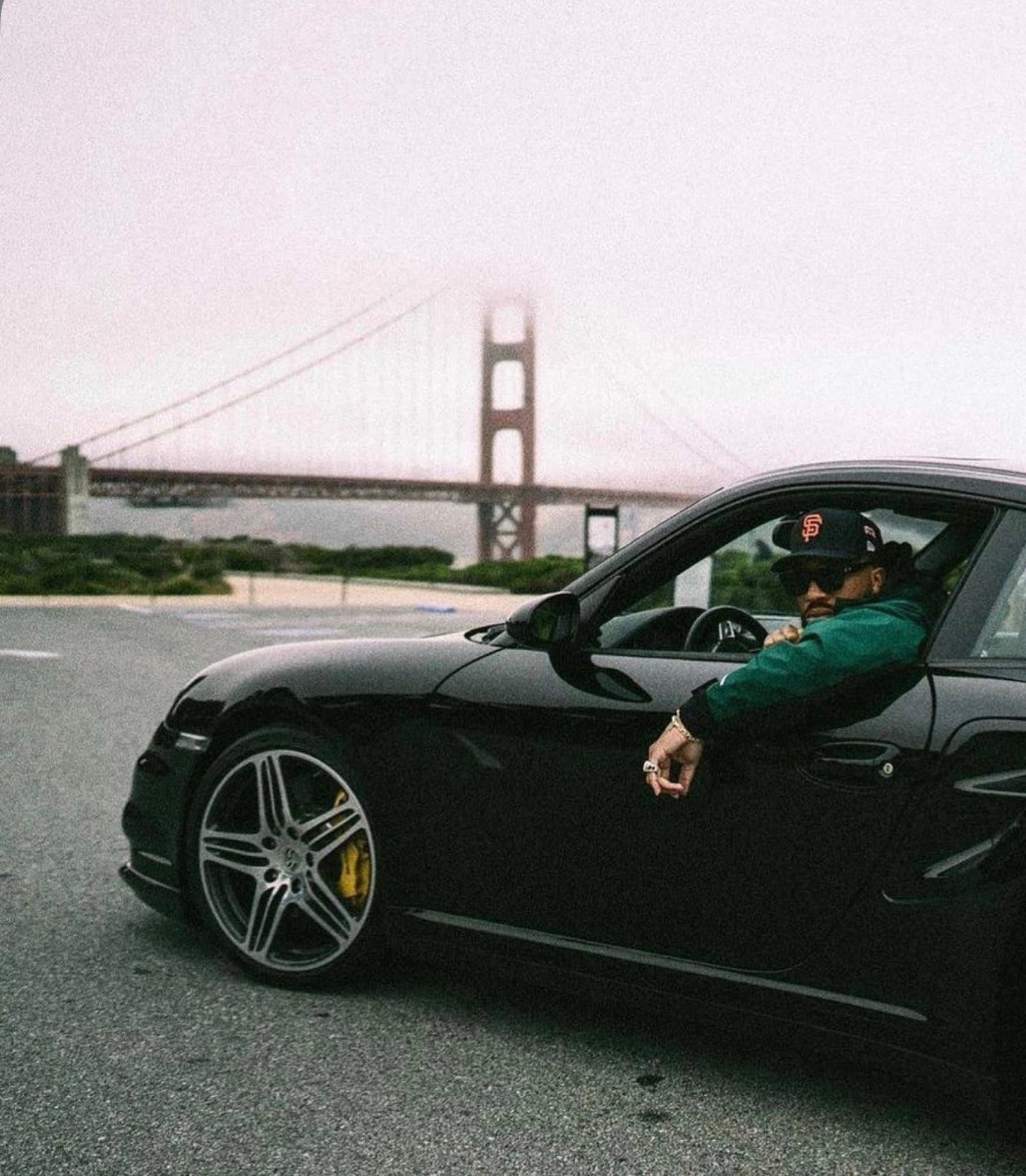 A man in a San Francisco Giants cap and green jacket leaning out the window of a black Porsche 911 (997 Turbo) with the foggy Golden Gate Bridge behind him.
