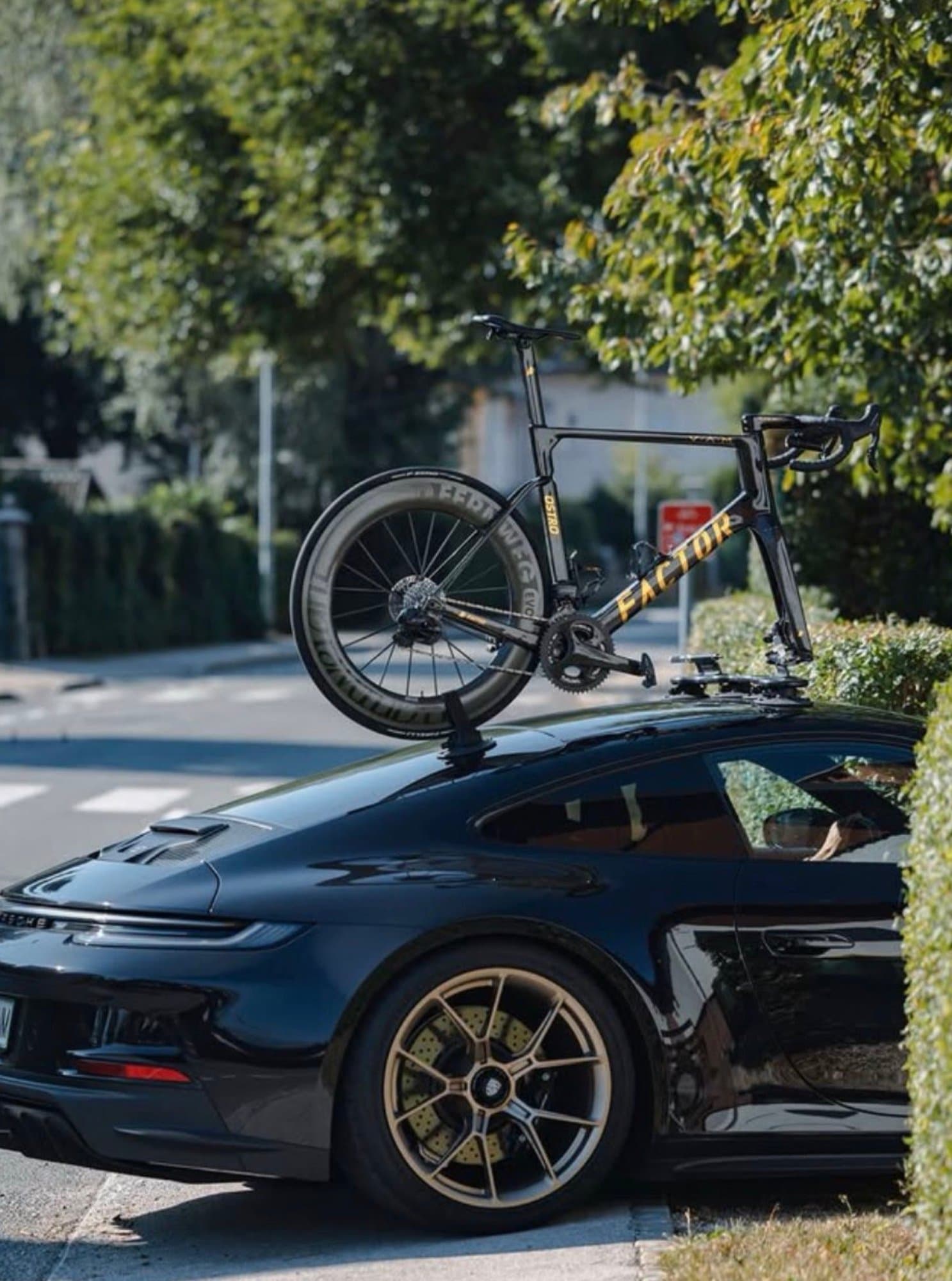 A gloss black Porsche 911 fitted with bronze wheels carries a black and gold Factor Ostro VAM road bike on a roof-mounted suction rack.