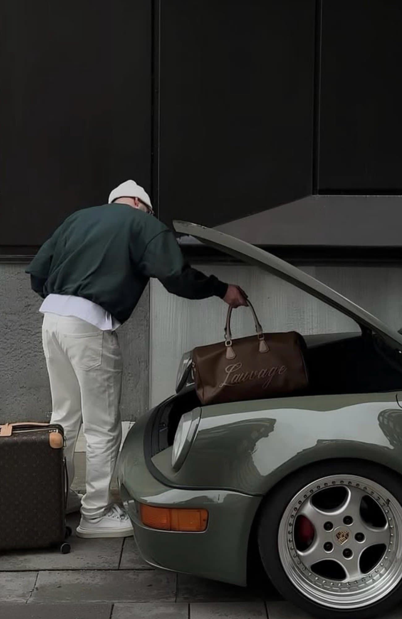 A man loading a brown leather duffle bag into the front trunk of an olive green vintage Porsche 911.