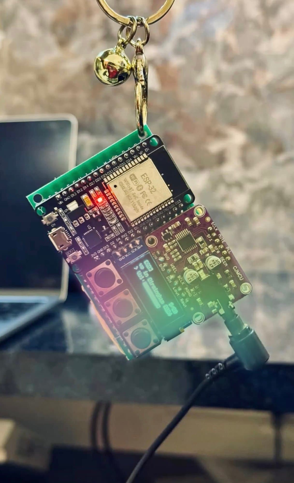 A DIY ESP32 digital audio player made of exposed circuit boards and a small OLED screen, suspended from a golden keychain with a bell.