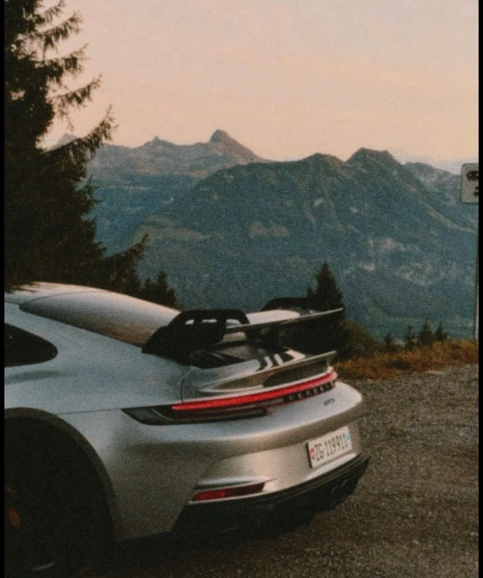 Rear three-quarter view of a silver Porsche 911 GT3 (992) parked along an alpine mountain road at dusk.