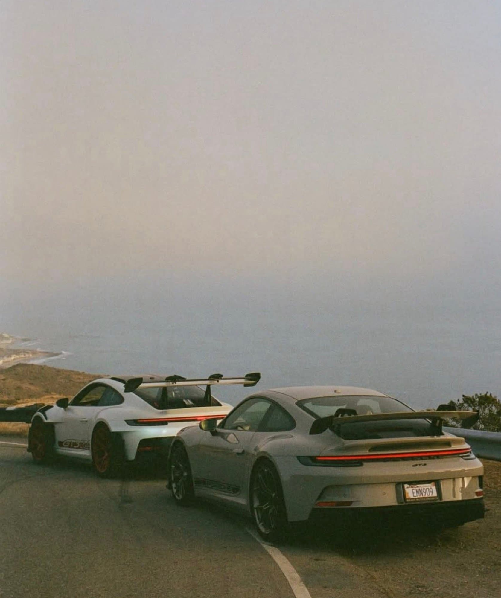 Two Porsche 911 GT3 (992 generation) sports cars parked alongside a coastal road overlooking a hazy ocean.