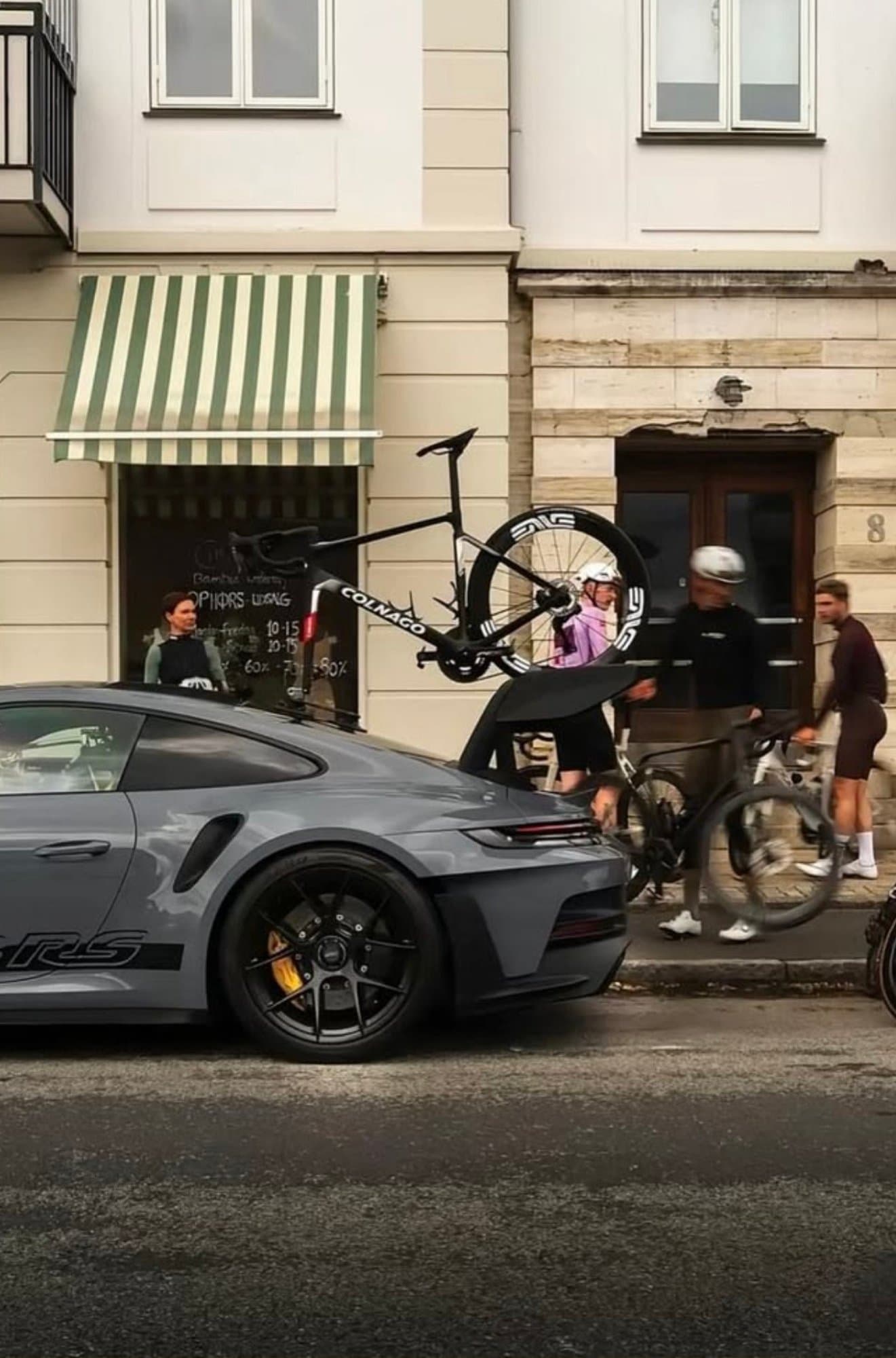 A Porsche 911 GT3 RS finished in slate grey parked on an urban street with a Colnago road bicycle mounted to its rear window suction rack.