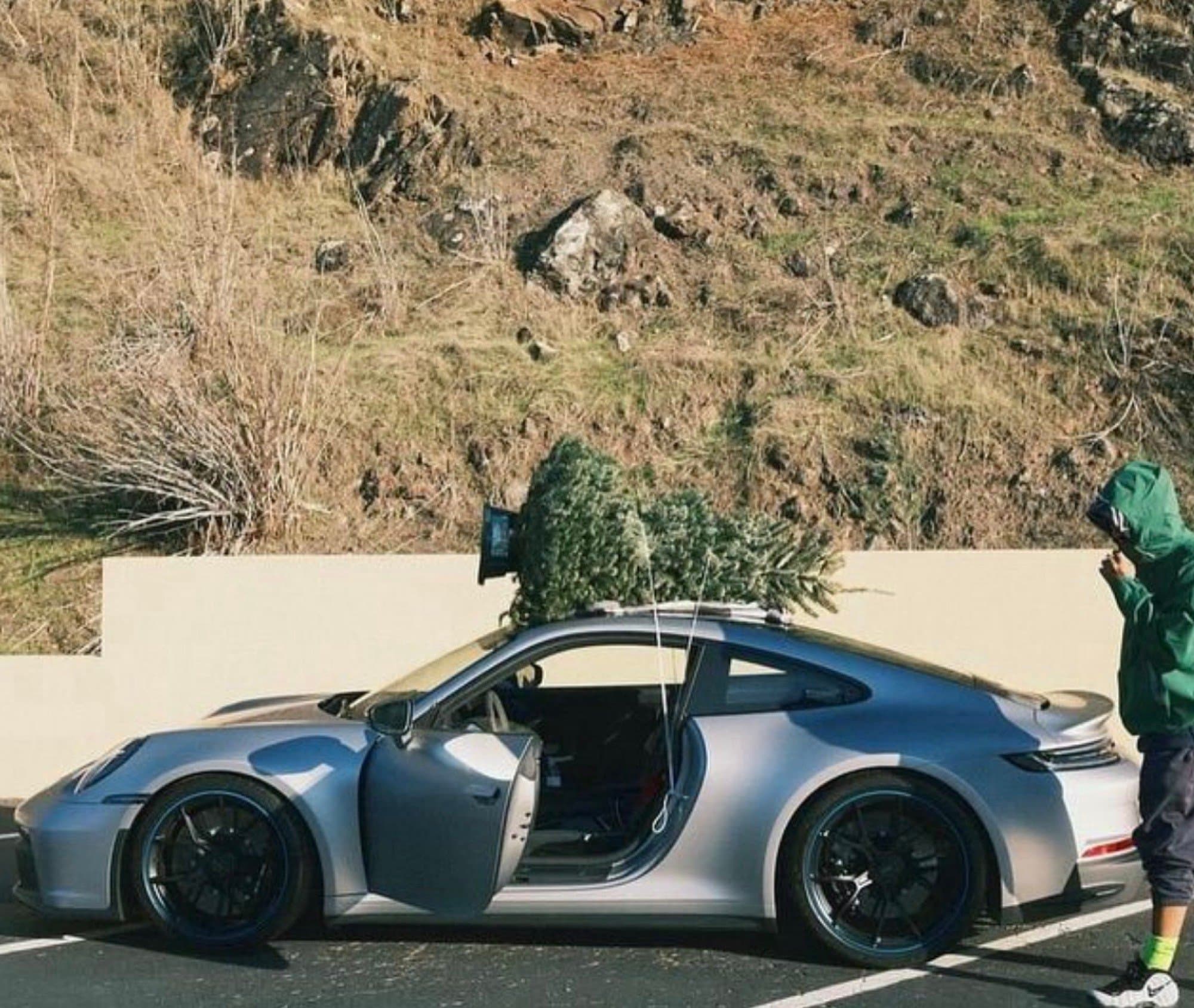 A silver Porsche 911 (992) parked against a rocky hillside with a freshly cut Christmas tree strapped to its roof.