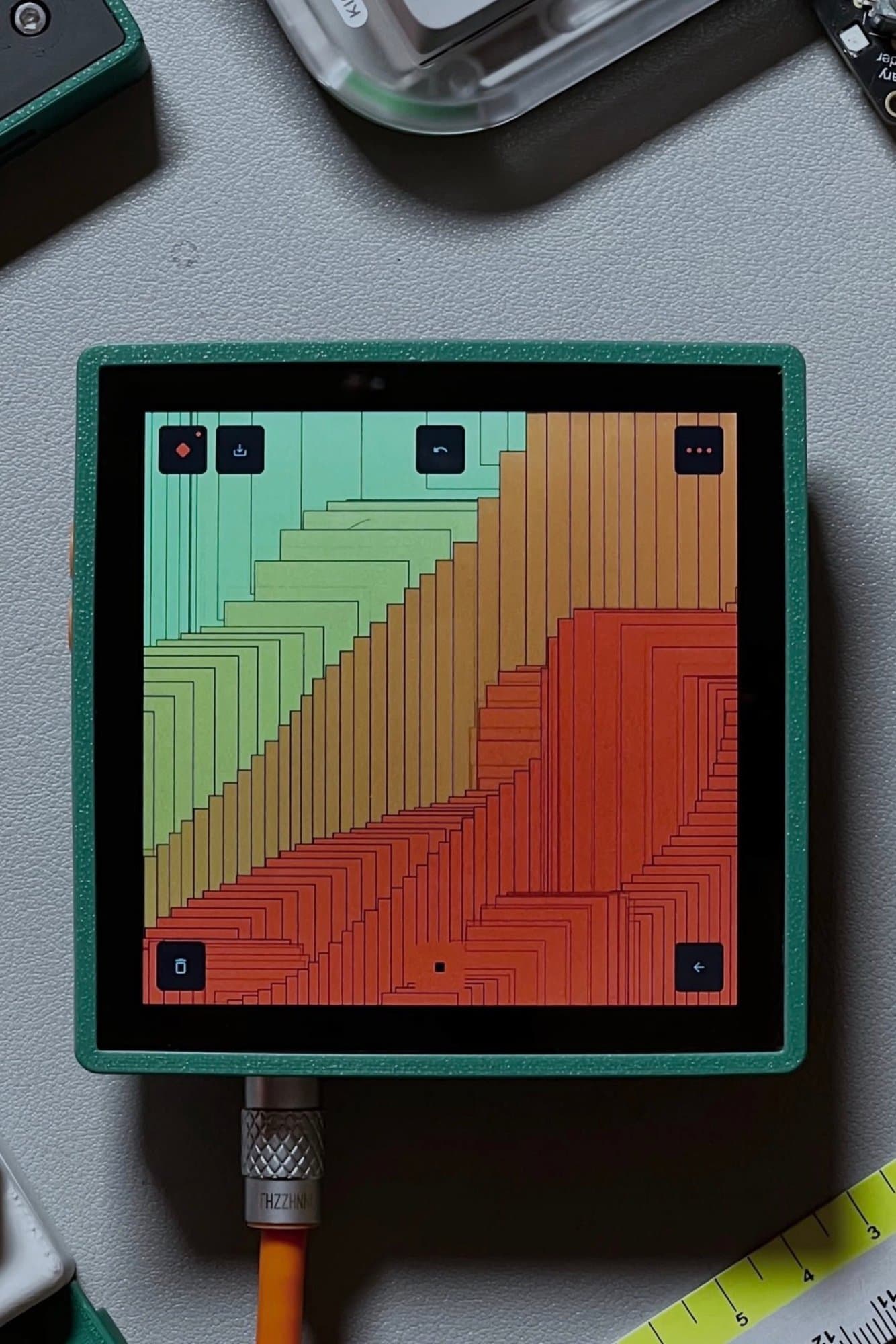 A compact square electronic display encased in a textured green frame showcasing generative geometric artwork.